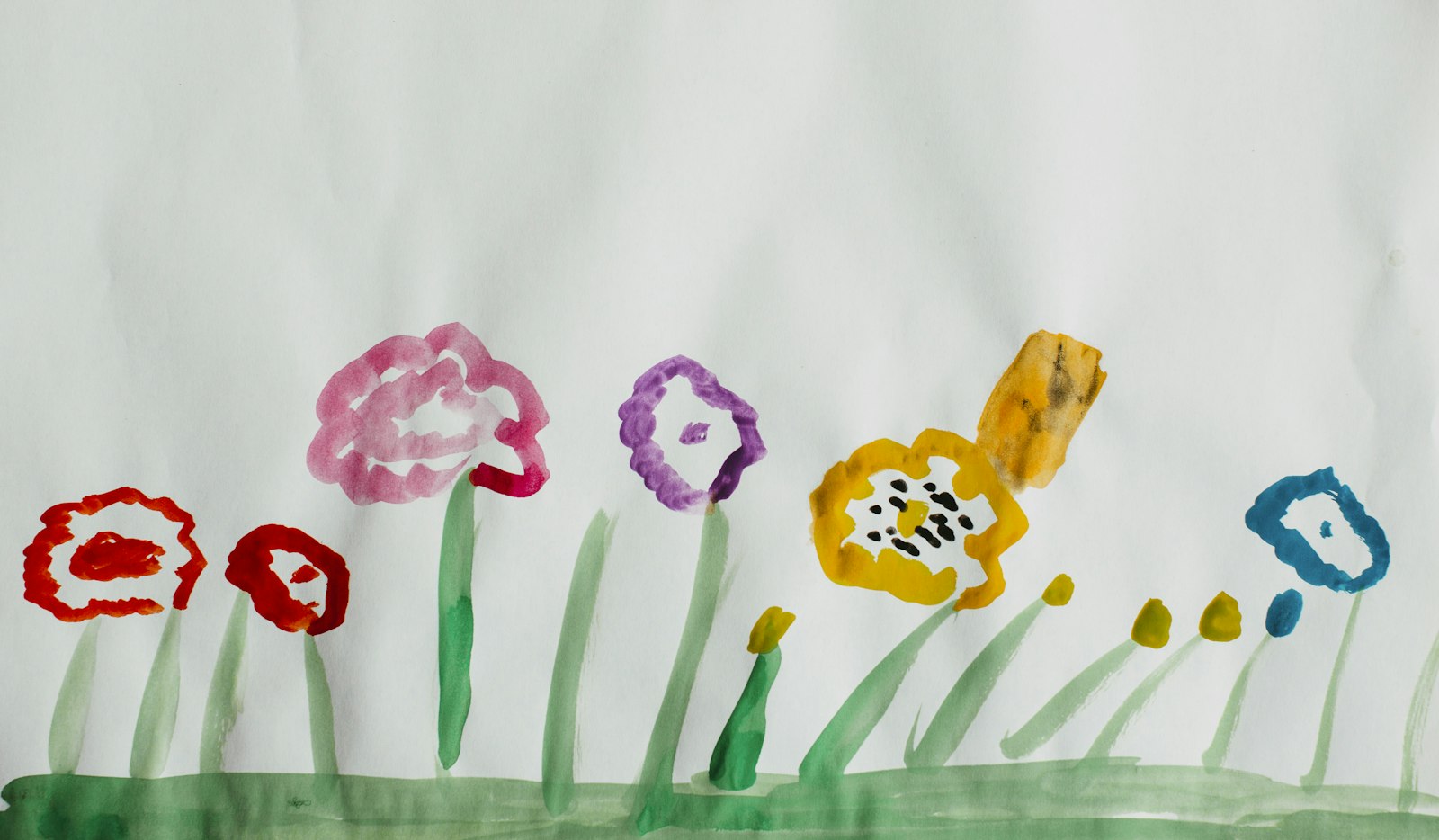 Un dessin d’enfant à la peinture : des fleurs rouges, rose, violette, jaune et bleue sur une bande d’herbe verte.